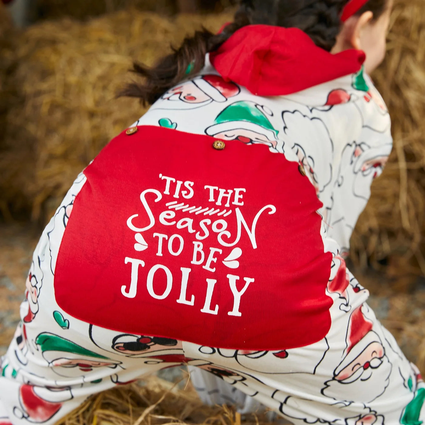 kid wearing a santa claus onesie with 'Tis the Season to be Jolly text, standing on hay.
