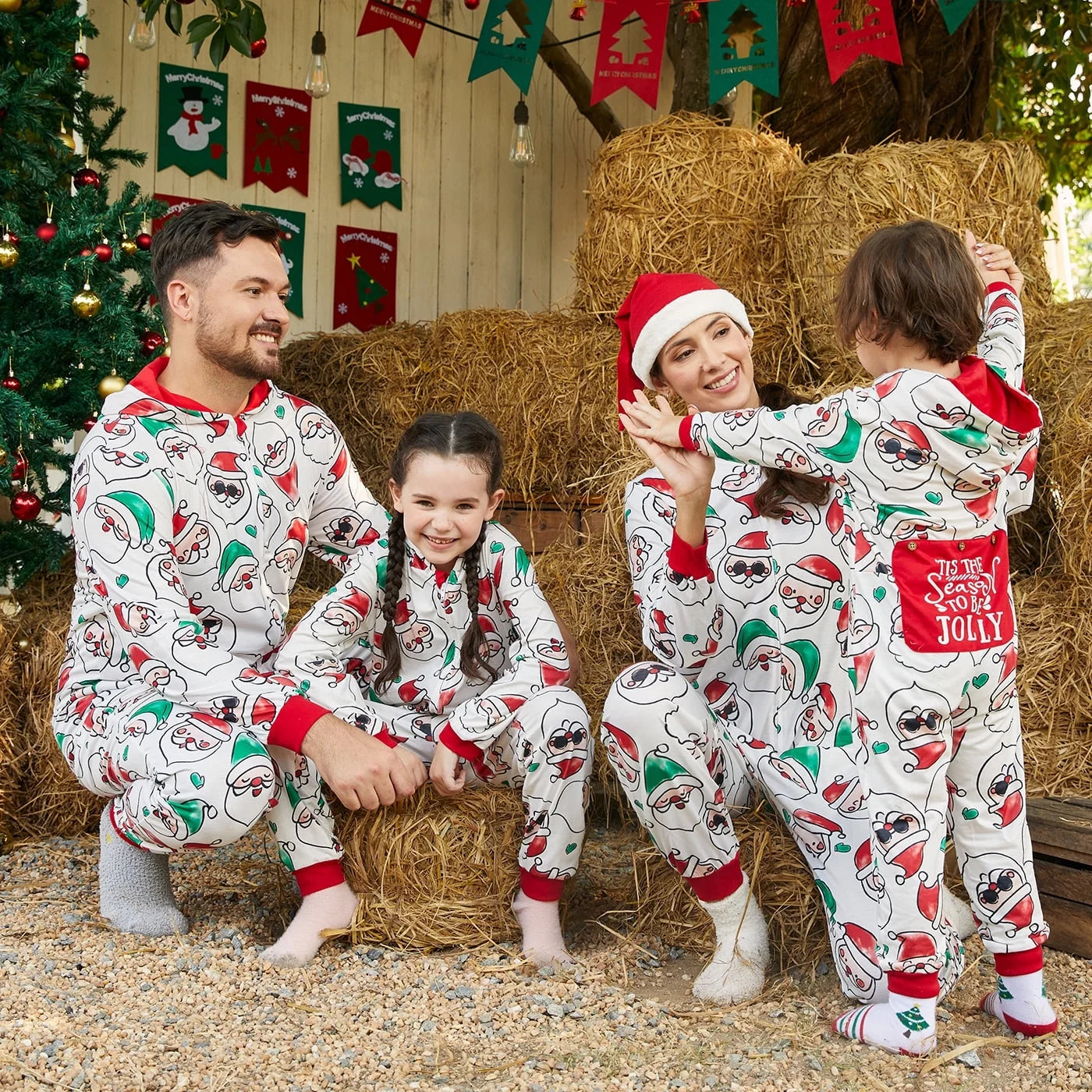 Family in matching Christmas pajamas playing outdoors with a decorated tree and hay bales in the background.